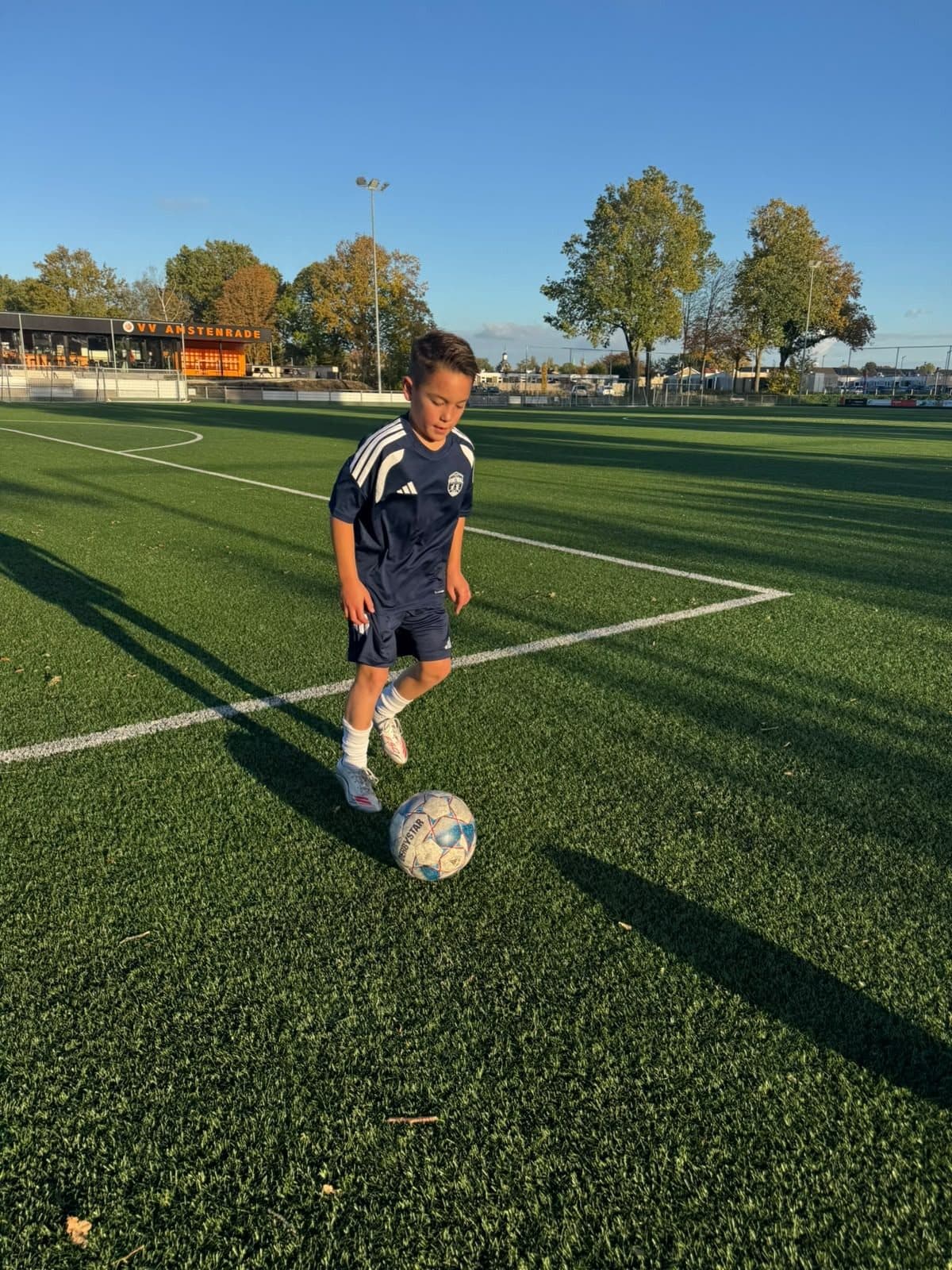 Een jongen in Voetbalfabriek-tenue dribbelt in de herfstzon over het veld van VV Amstenrade